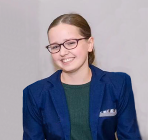 A white woman in a manual wheelchair smiles at the camera. She is wearing glasses, a green shirt with a blue jacket, and has brown hair in a low ponytail.