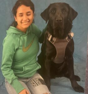 Headshot of Karlee Rojas, an olive-skinned Latina woman with dark brown hair wearing a green sweatshirt and jeans seated next to her service dog, a black lab.