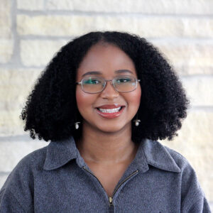 Alana's head is centered against a white back ground. She's wearing a grey jacket and glasses. Her hair is down and she's smiling.