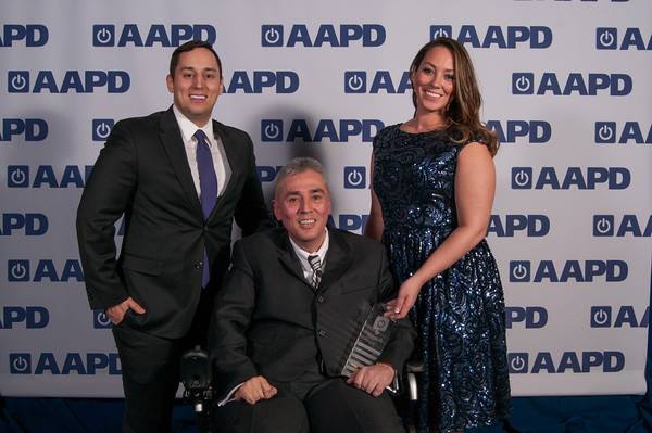 An image of three people smiling in front of a white background with the AAPD logo. In the middle, Andrés Gallegos, a Latino man with grey salt-and-pepper hair is wearing a black suit and holds an award in his right hand. He is smiling at the camera. To his left is his son, a Latino man with black short hair smiling at the camera and wearing a suit. To his left is a woman with tan skin and brown hair waring a nice blue dress