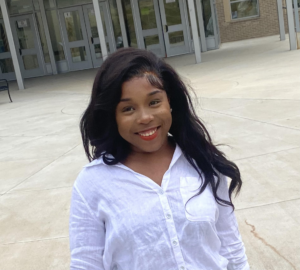 Kaysia, a black woman with long hair passing her shoulders is wearing a white shirt and smiling towards the camera