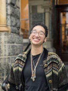 Ollie, a short haired fair-skinned person with glasses, smiles in front of a stone building. They're wearing an open green sweater with three crochet necklaces and small green pendant earrings.