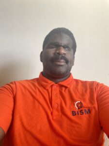 The picture is a close-up selfie of a man. He has dark skin and short black hair. He is wearing a bright orange polo shirt with a collar. On the left side of the shirt, there is an emblem of an eye inside a circle, and next to it, the letters 
