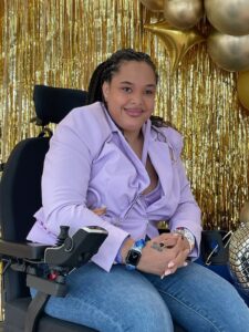 Saphire Murphy is medium tone bi- racial woman in her late 20’s with box braids, a lavender blazer and top and blue jean jeans in a power chair. She smiling at the camera.