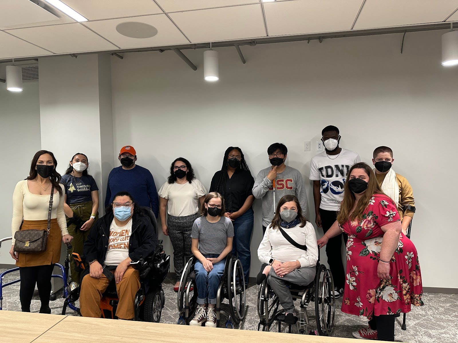 A group of twelve diverse individuals posing for a photo indoors. Several people are wearing face masks, and three individuals are seated in wheelchairs.