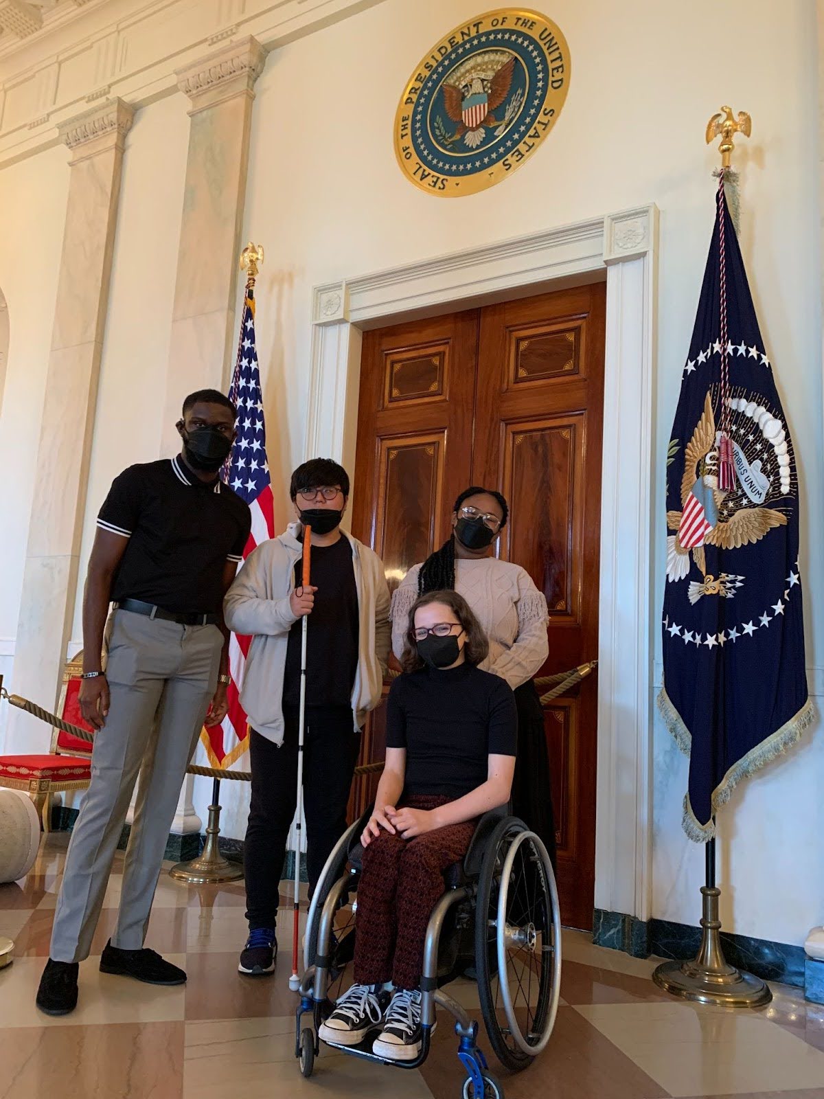 Four people stand in front of the Seal of the President of the United States, between two American flags. One individual is using a white cane, and another is seated in a wheelchair, all are wearing masks.