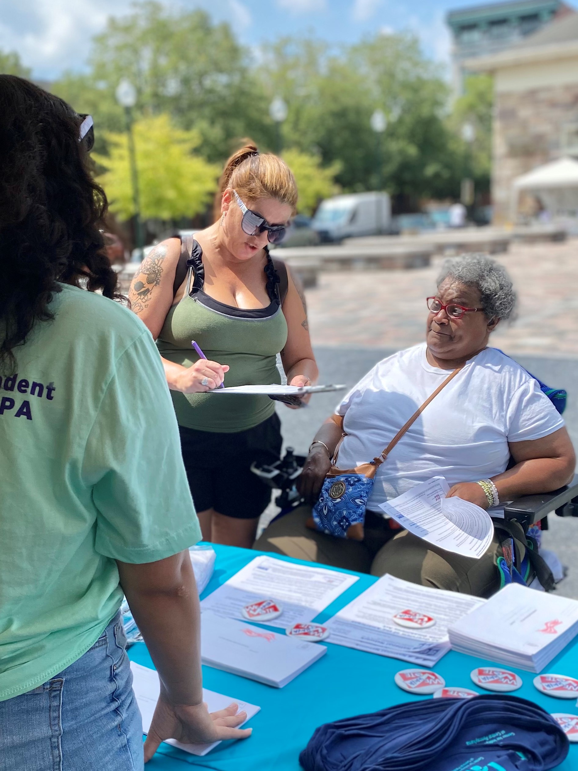 Three people at an outdoor information booth, with a woman writing on a clipboard and an elderly lady in a wheelchair.