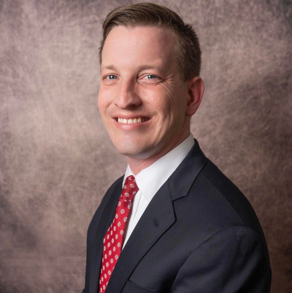 Michael is a white man, smiling over his left shoulder. He's wearing a dark blue suite, white collared shirt, and red tie. 