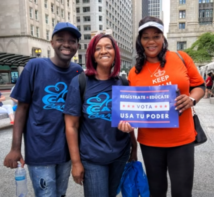 Three smiling people, two wearing blue shirts with 'Care' logos, and one in an orange shirt, holding a 'Register, Educate, Vote' sign in Spanish.