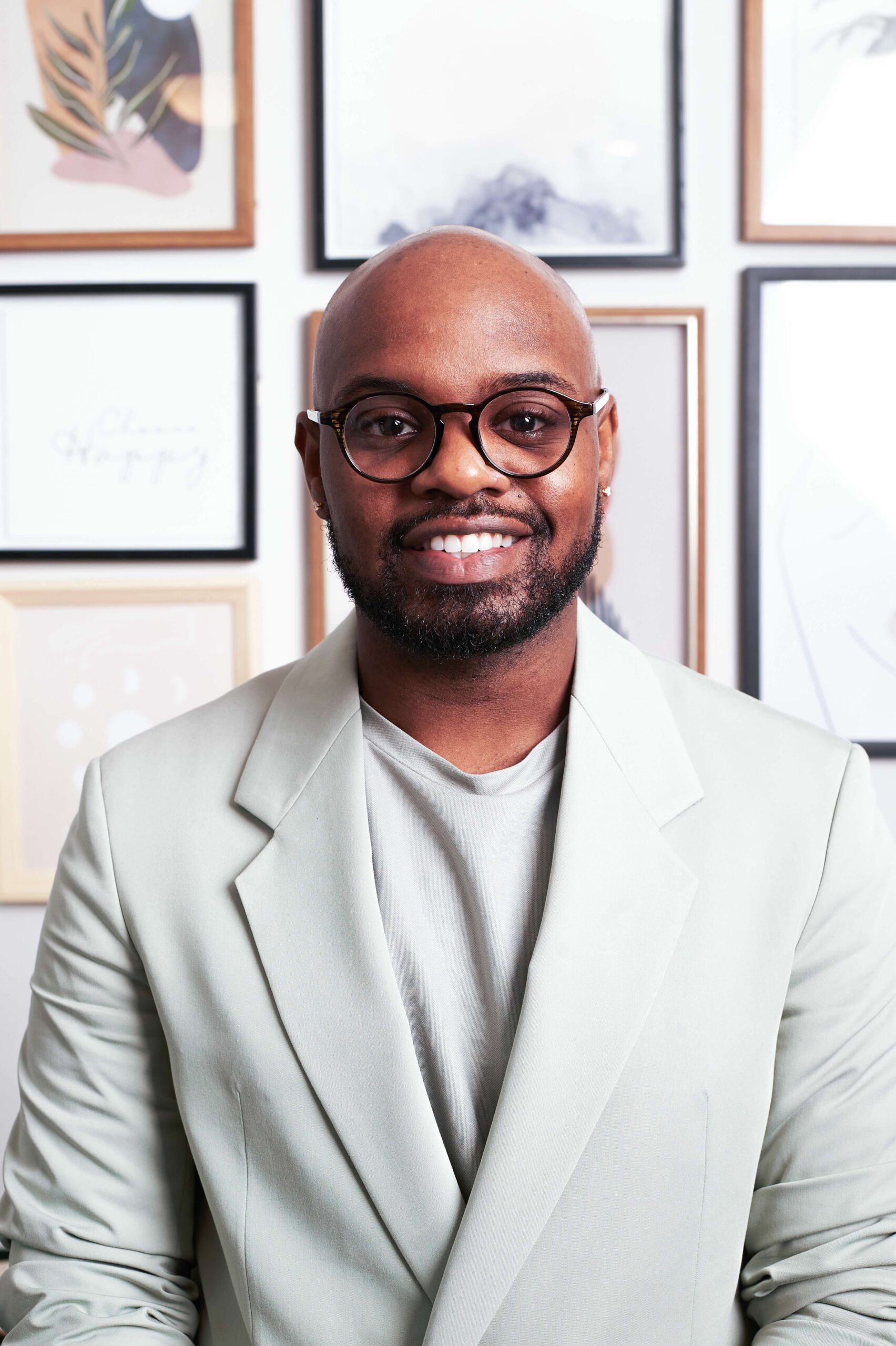 Dewayne Johnson (he/they), a Black person with a bald head and neatly trimmed beard, smiles directly at the camera. He is wearing a light grey suit and shirt that are stylish and monochrome, and black round glasses. Behind them is a gallery wall with posters and art.