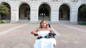 Headshot of Fabiola, a brown woman sitting on a black and pink wheelchair. Fabiola has medium length blonde hair that is curled. She is wearing a white dress and looking at the camera and smiling. Behind her is part of the UT tower which consists of white bricks and concrete.
