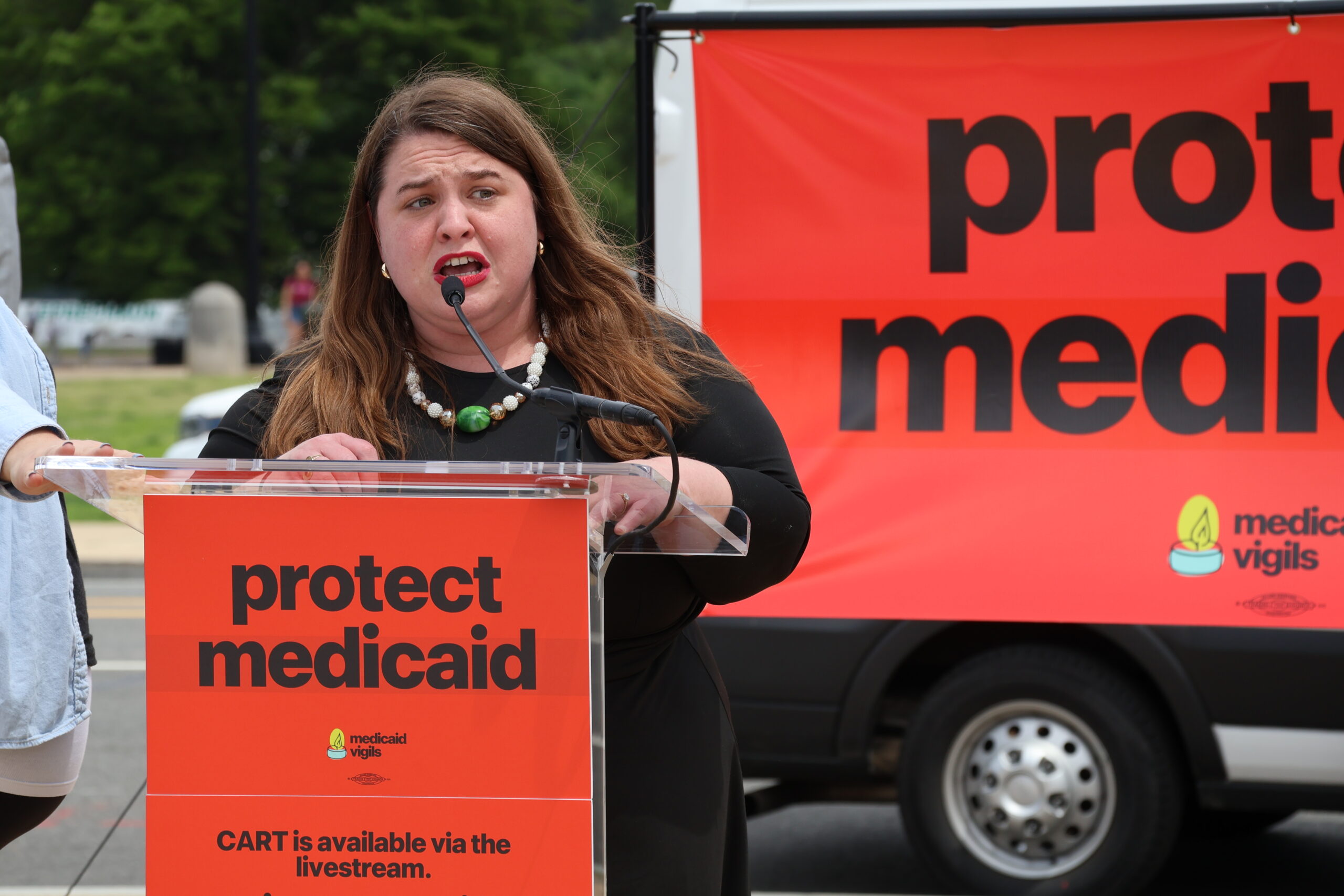 IMG_0176 Image of AAPD President and CEO Maria Town, a white woman with long brown hair and red lipstick, standing at a podium with a "protect medicaid" sign on it. She is speaking at the 24 hour vigil to protect Medicaid.