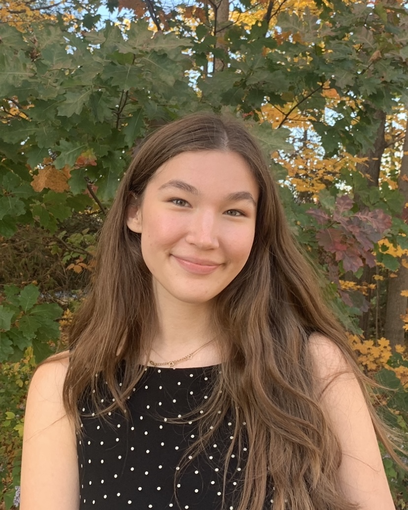 Aubrianna Wilson, a mixed race (White and Japanese) young woman with long brown hair, is smiling on a bright autumn day. She wears a black and white polka dot top.