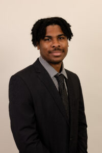 ID: Black man with short twisted hair, a dark black suit, and a black and white collared shirt and tie.