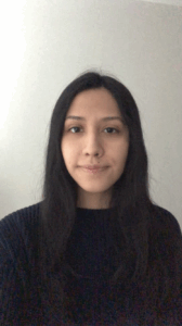 A portrait of a Latinx person with brown eyes and long dark hair. They are facing forward and are framed in the middle of the photo. They are wearing a navy blue knit sweater and they are in front of a light gray wall.