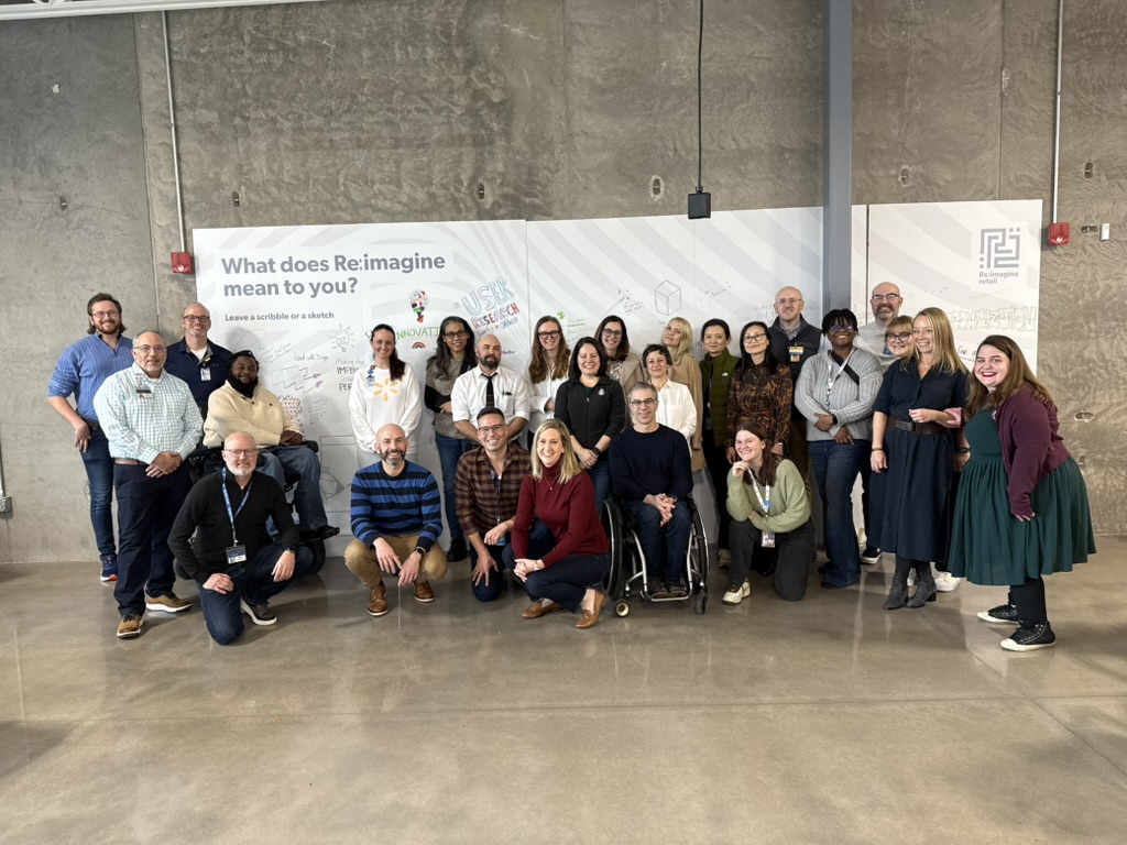 A group of seven people, six sitting and one standing behind them, posing for a photo at a long wooden table with microphones and Starbucks cups in front of them. The setting appears to be a modern, well-lit room with large windows and a screen on the wall in the background. Everyone is smiling and dressed in casual or business-casual attire.