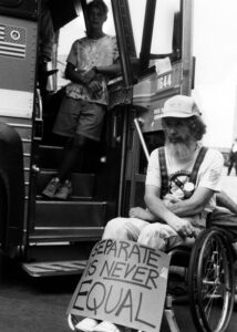 A black and white photo of Bob Kafka, a white man with a long beard, protesting outside of a bus in his wheelchair with a sign that says "Separate is NEVER equal" 