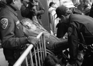 Bob Kafka, an older white man who is a wheelchair user and has curly white hair and a long white beard, getting arrested while in his wheelchair