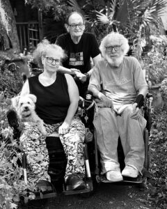 Bob, an older white man with curly white hair and and a beard with his with Stephanie, who is also a wheelchair user and has a little white dog on her lap, and Tom Olin standing behind them