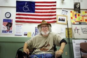 Bob, an older white man with curly white hair (under a hat) and a beard sits in his wheelchair and smiles at the camera. He has the ADAPT flag and a REV UP poster behind him. 