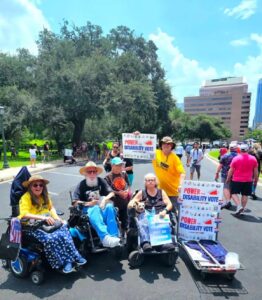 A number of individuals in wheelchairs and standing at a REV UP event. they are holding protest signs and American flags. Bob Kafka is in the group, smiling