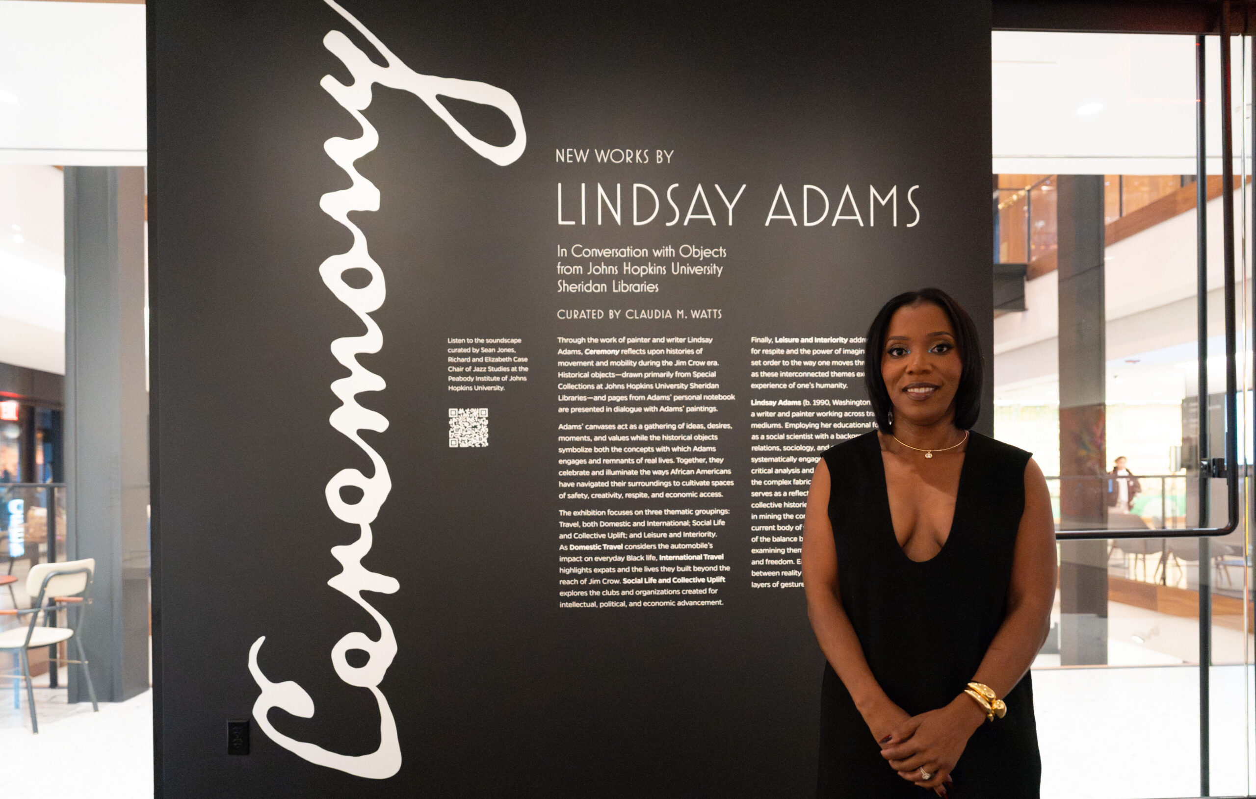 Lindsay Adams standing in front of a wall detailing her exhibit called “Ceremony.” She’s wearing a black dress and gold jewelry.