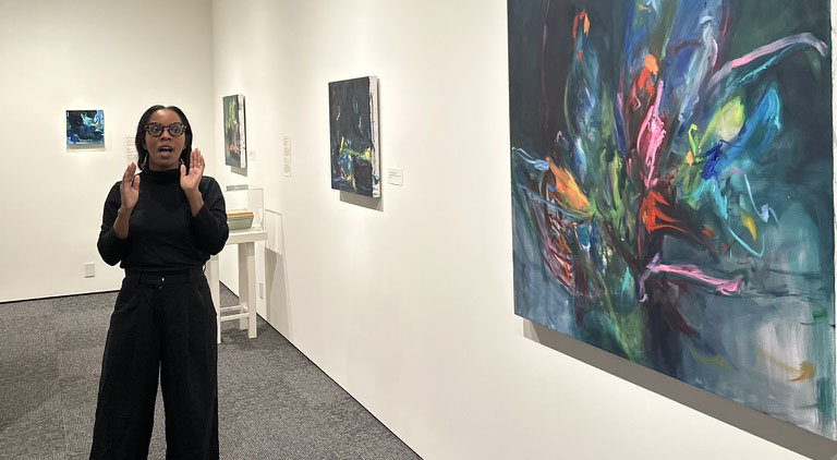 ID: Lindsay Adams speaks next to one of her paintings called “Lotus Blossom”. The painting features many colors on a primarily blue background. The painting looks like a lotus flower blossoming.