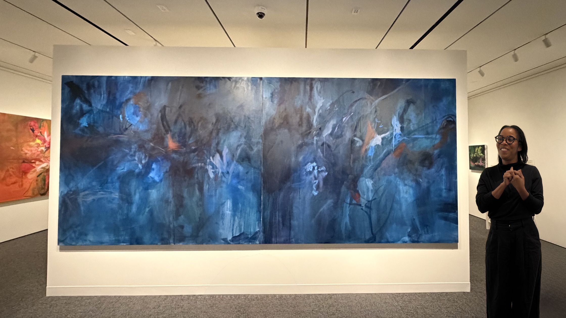 Lindsay Adams smiles while speaking next to one of her paintings called “Kind of Blue.” The painting is large and takes up the entire wall that it is on. The painting features a blue and black motif, with small highlights of red throughout.