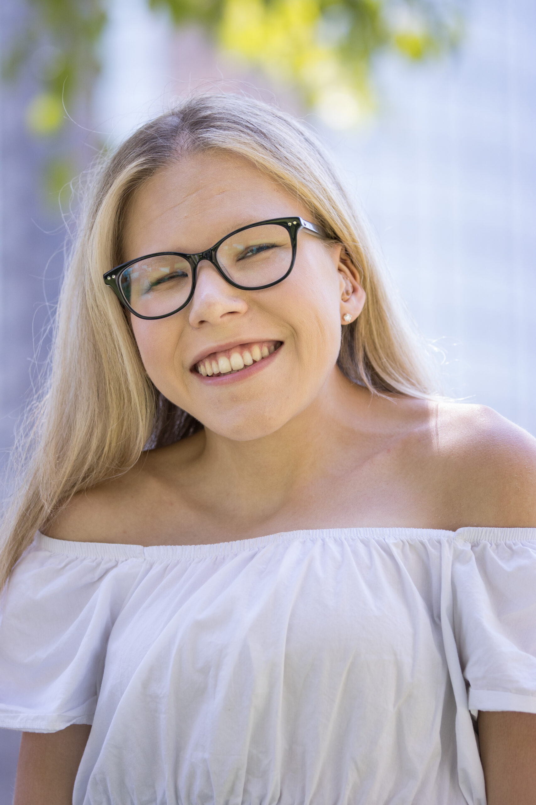 A smiling blonde woman wearing black-framed glasses and a whiteoff-the-shoulder top stands outdoors in soft natural light, with greenery blurred in the background