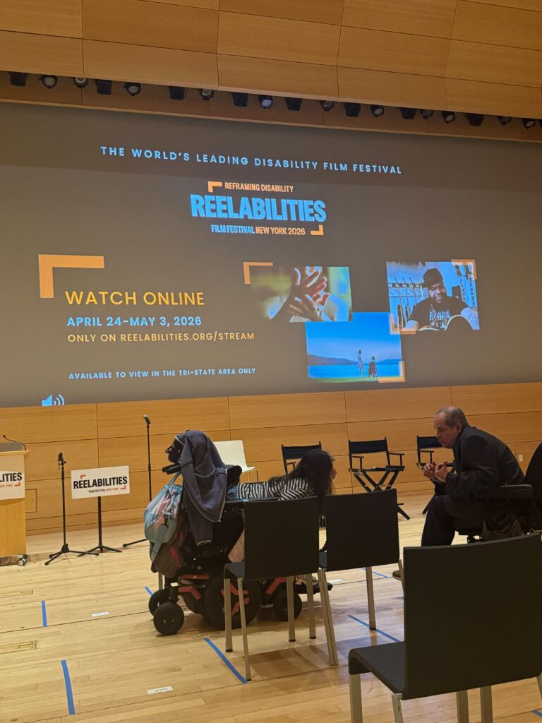 Two wheelchair users speak, in front of a projected ReelAbilities sign.