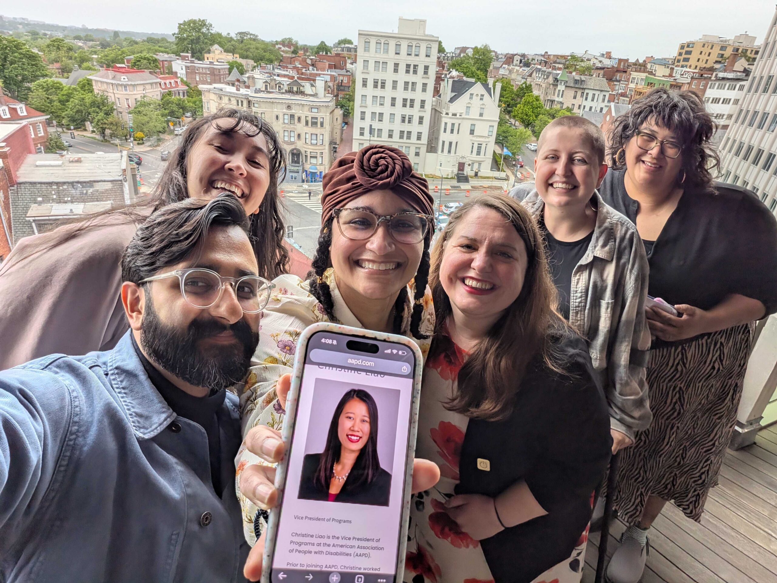 A group selfie of staff taken on the rooftop of the AAPD alumni event venue. From left to right, Neil, April, Sarah, Maria, Katie, and Gabi smile towards the camera, as Sarah holds up a photograph of Christine, who was unable to make the event.