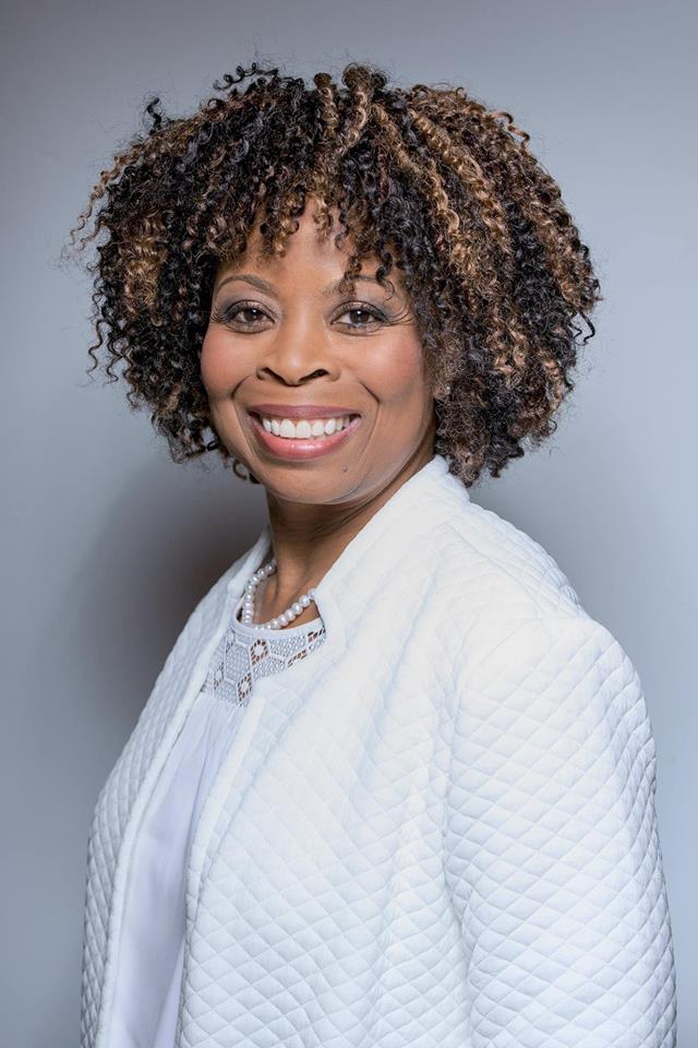 Headshot of a woman with curly hair wearing a white blazer, smiling against a gray background.