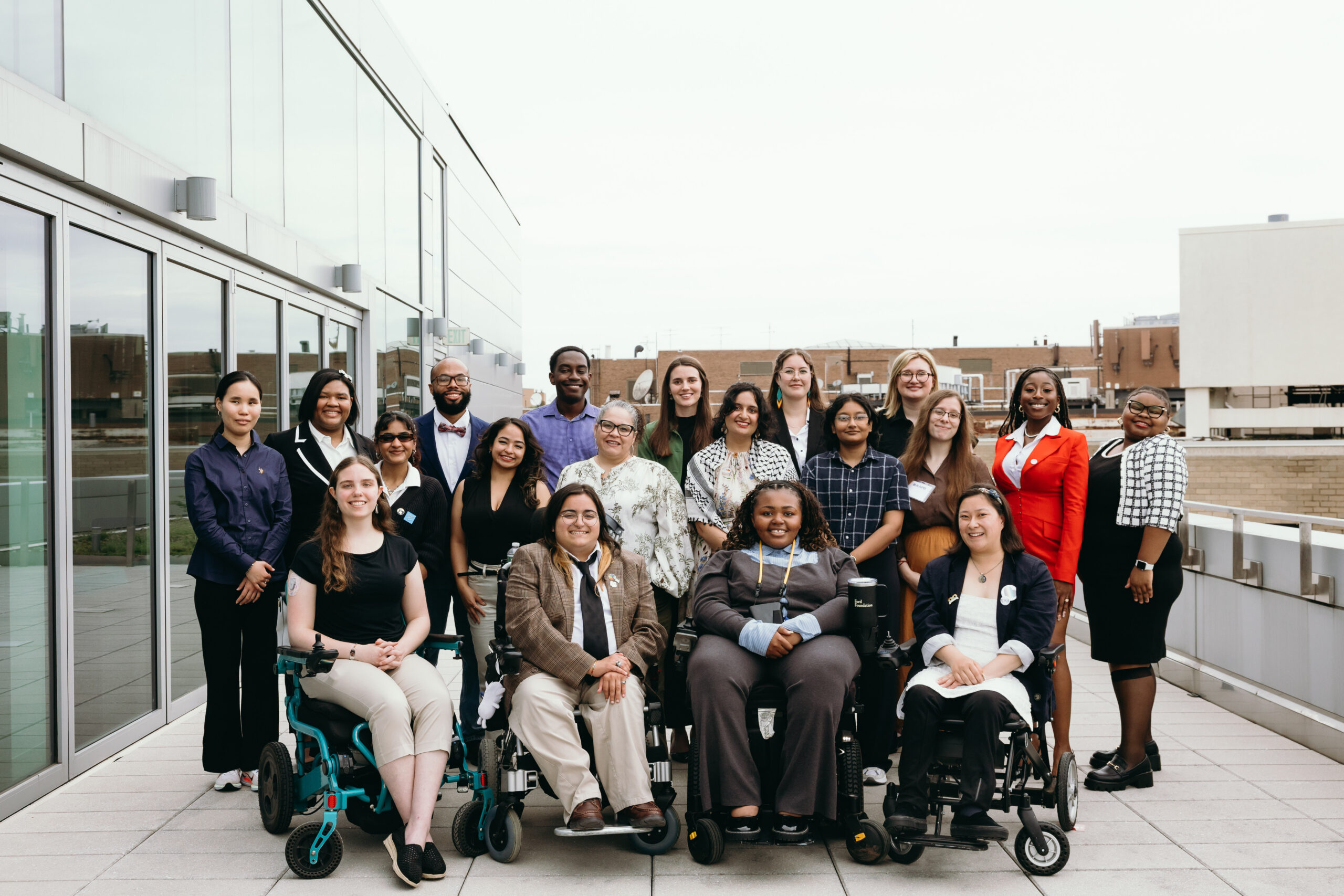 A group of AAPD interns smile at the camera, gathered outside on a rooftop balcony. The group is a diversity of races and genders, and has a diversity of disabilities. Some members of the group are sitting in wheelchairs, while others are standing.