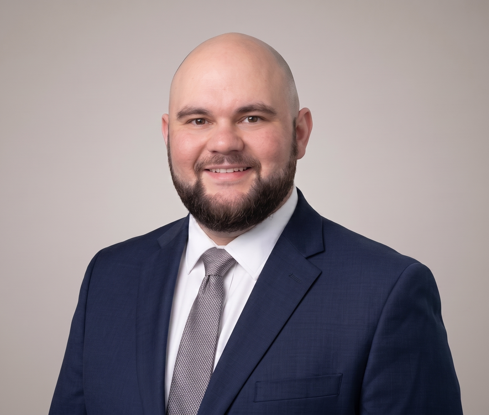 Headshot of a smiling, bald man with a beard wearing a navy blue suit jacket and a grey tie against a neutral studio background.