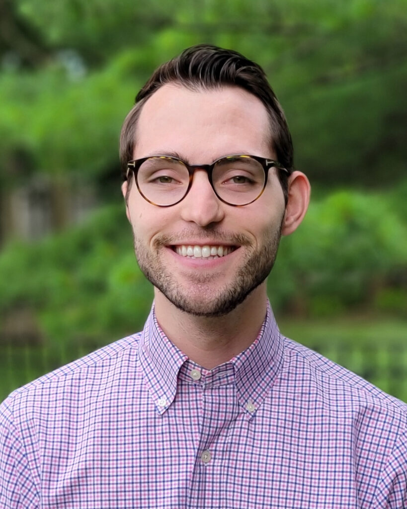 A photo of Jack Reeves in a business-casual button-down smiling at the camera in front of trees
