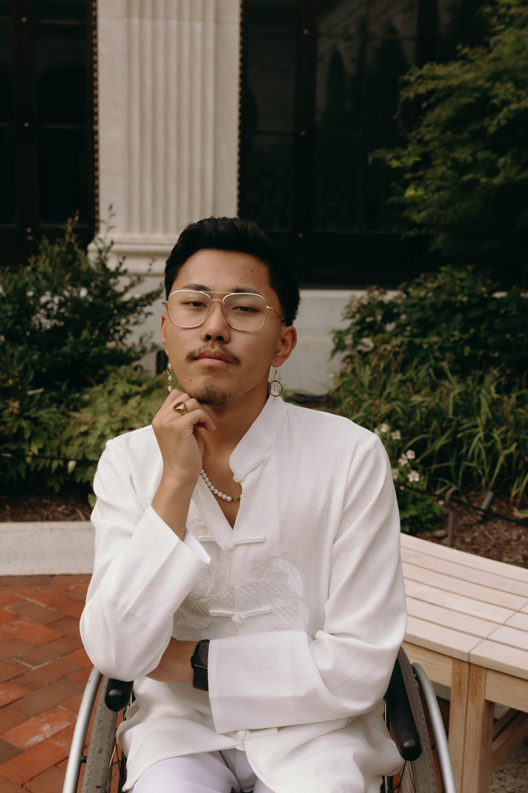 HuáHuī, a Chinese person in their early 20s with black hair, is sitting in their wheelchair, with their right arm over their body and left arm folded under their chin. A face postured in deep thought. They are wearing a white embroidered dragon button-down. They have round-shaped gold glasses, a pearl necklace with a chain, and gold-pearled earrings.