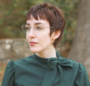A headshot of KD, a white woman with short brown hair and clear glasses wearing a long sleeve emerald green blouse with a bow near her neck. KD is looking away and smiling softly.