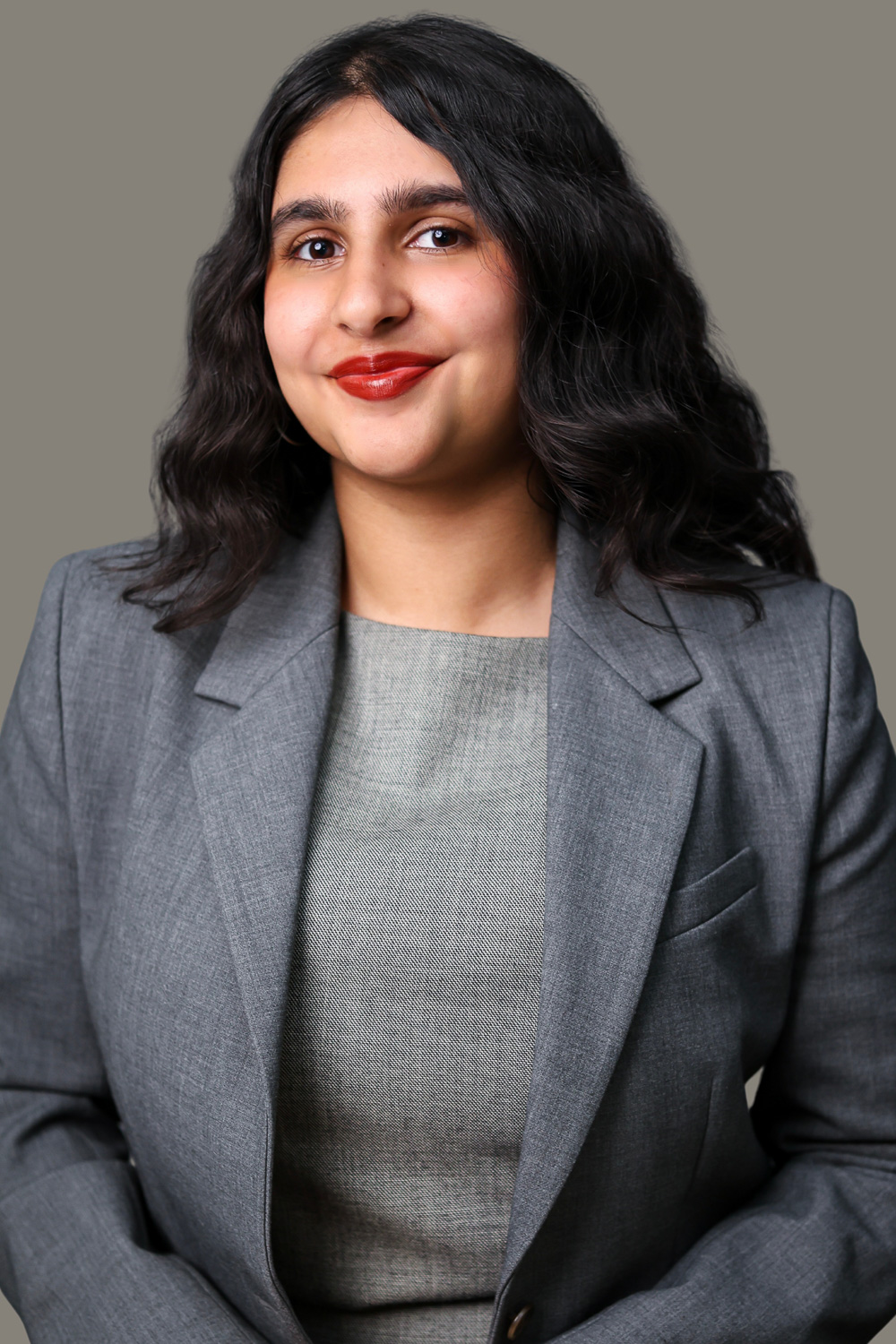 A studio headshot of a warm-toned South Asian person. They have long dark hair parted in the center. They are dressed in a grey blazer over a light grey blouse. They are facing the camera, head slightly turned to one side, with a close-lipped, friendly smile.