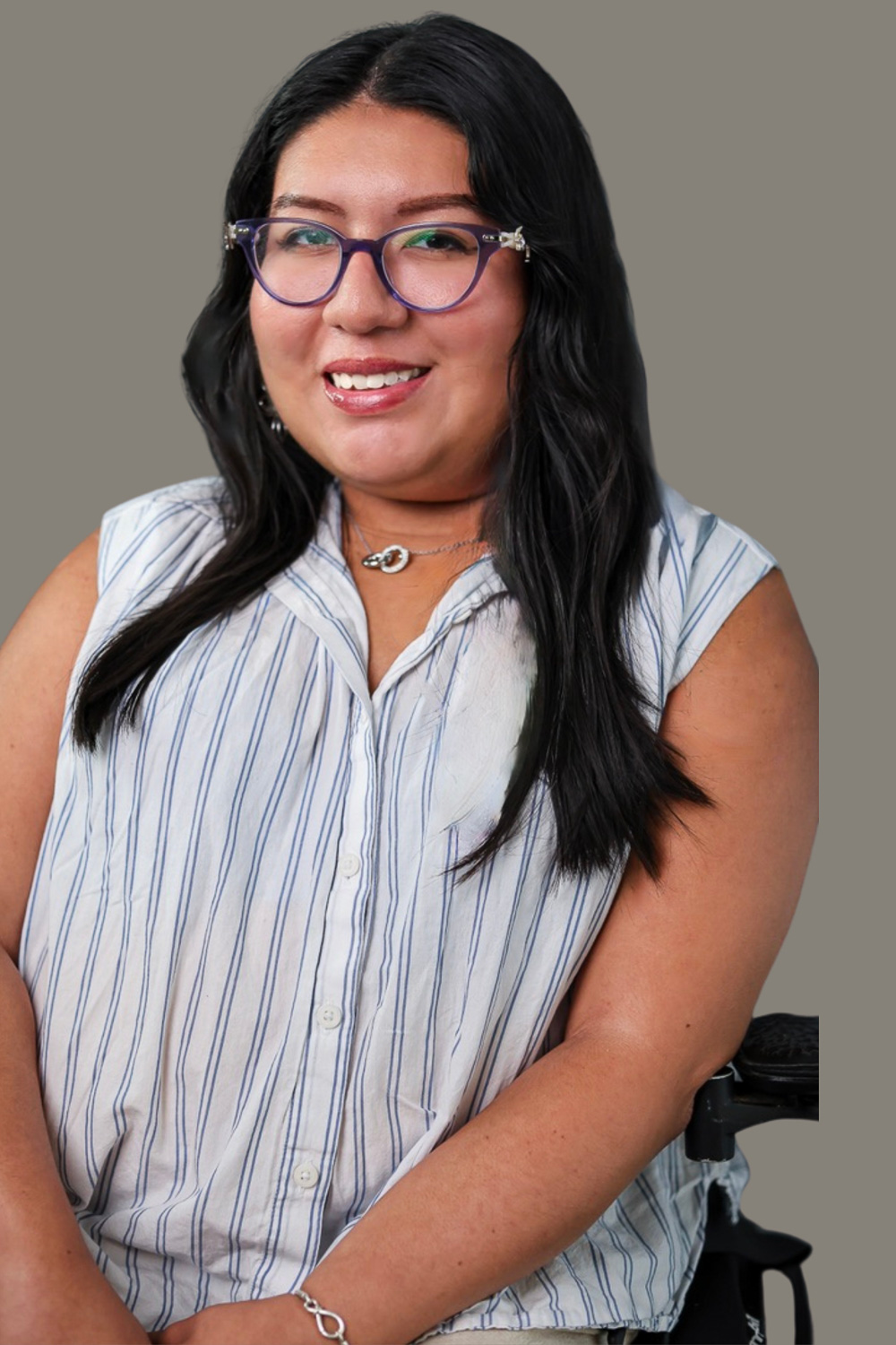 A warm toned Hispanic woman with long black hair and purple glasses. She is smiling and wearing a glossy pink lipstick. Maria is wearing a sleeveless buttoned up white dress shirt with blue stripes. She also wears a silver necklace of two intertwined rings and a silver infinity bracelet.