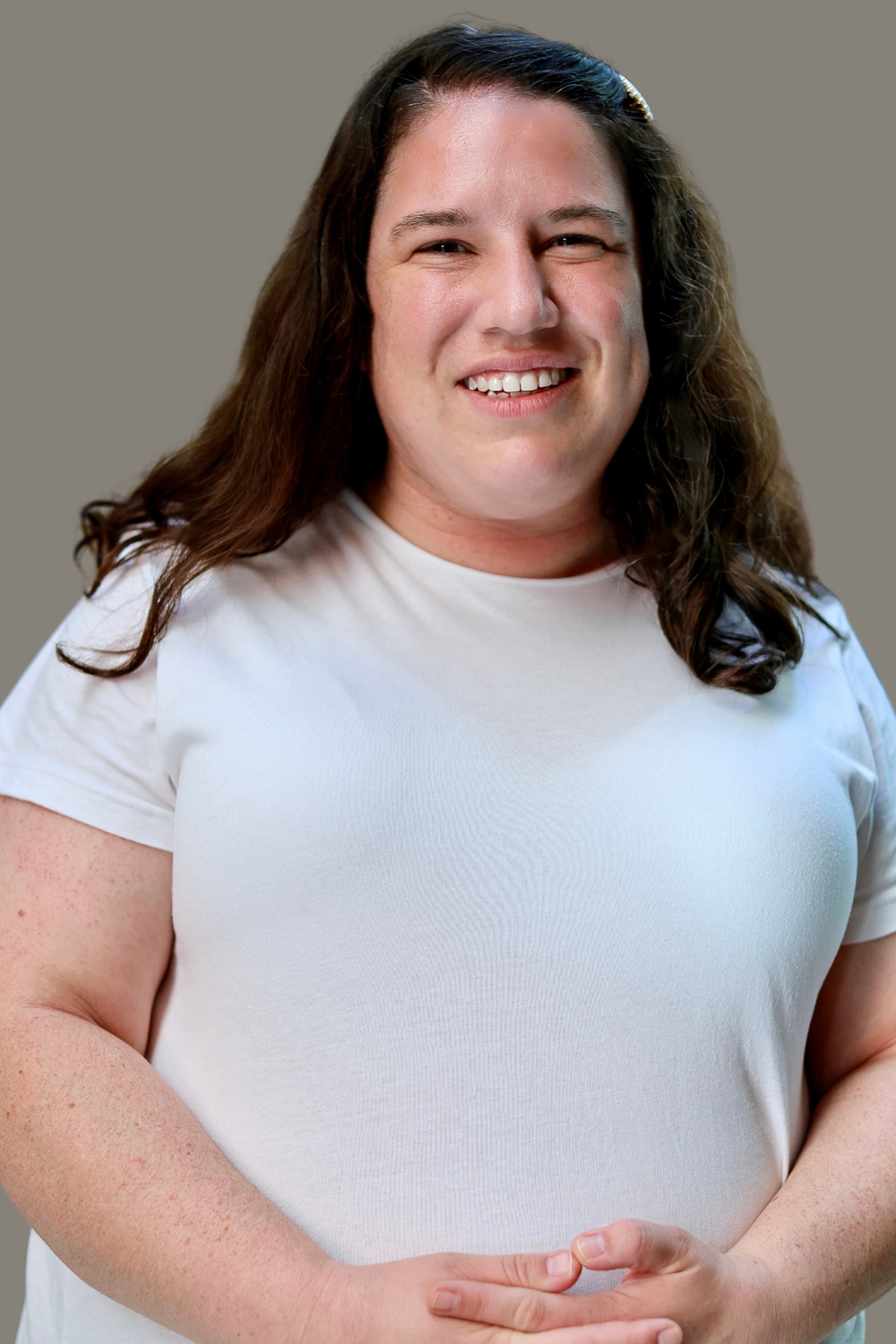 Tori is a white woman standing in front of a beige background. She has shoulder length, wavy, dark brown hair and is wearing a white short sleeved shirt. Her hands are clasped together at her waist and she is smiling at the camera.