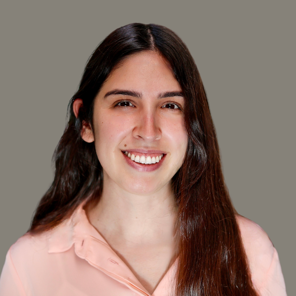 A light-skinned Latina with long brown hair is wearing a coral pink long-sleeved shirt and is smiling a toothy grin in front of a gray background.
