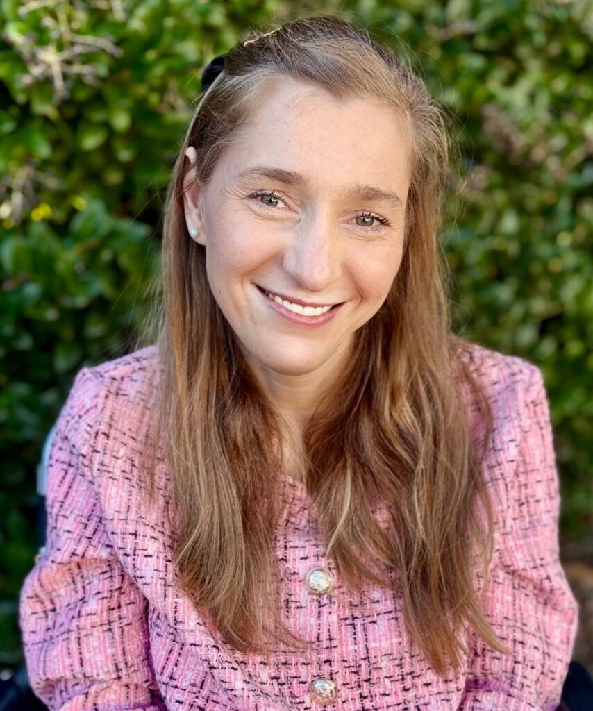 Photo of Lilly, a white woman with long dark blonde hair, sitting outside in front of leafy green foliage. She is wearing a pink tweed jacket with decorative buttons, pearl earrings, and a black bow in her hair. She looks directly at the camera and smiles warmly.