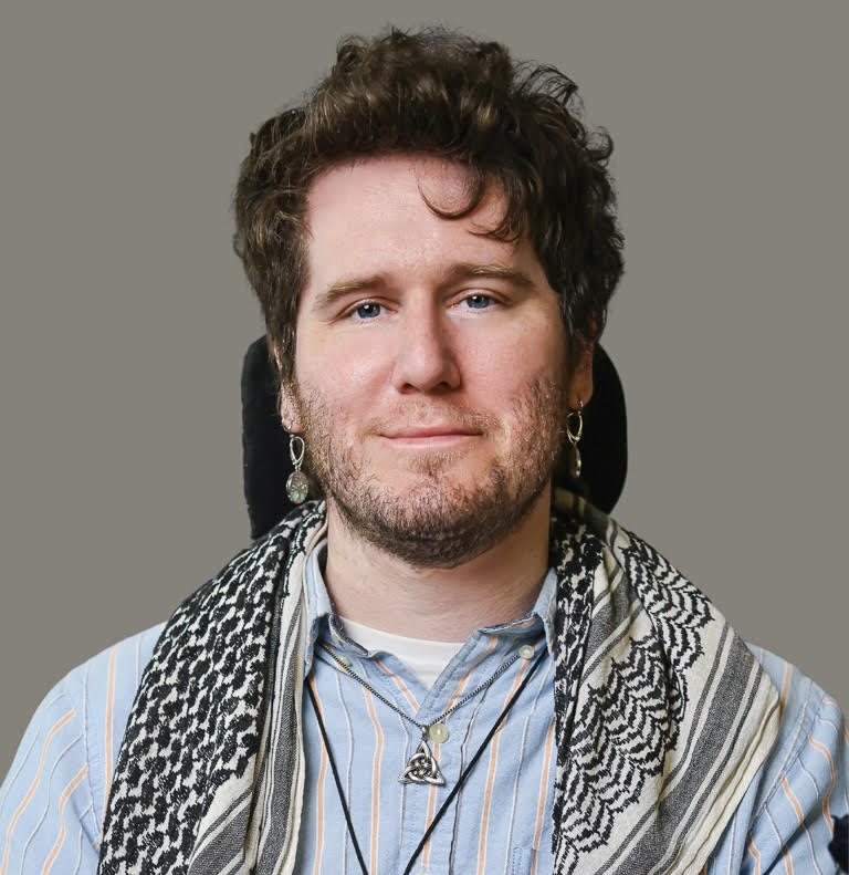 A headshot of Tre, a white person slightly smiling with wavy brown hair, a short beard, blue eyes, and silver and blue-green “tree of life” dangly earrings. They are wearing a periwinkle, coral, and gray vertically striped button-up shirt, a keffiyeh, and a silver necklace of a triquetra. In the background, there is the black headrest for their powerchair as well as a gray backdrop.