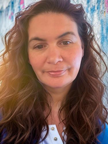 A person with long, wavy brown hair is shown in a close-up portrait, facing the camera with a calm, neutral expression. They are wearing a light blue top. The background is softly blurred with cool tones, creating a relaxed and approachable feel.